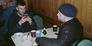 zwei Männer in einem Cafe, einer spielt mit dem Handy, vor sich Pappbecher