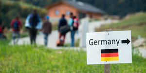 grüne Wiese, hinten laufen Leute auf einem Weg, vorne steht ein Schild mit Aufschrift "Deutschland" und nach rechts weisendem Pfeil