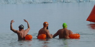 Drei Männer schwimmen zwischen orangenen Bojen im Gletscherwasser, im Hintergrund das Gletschereis