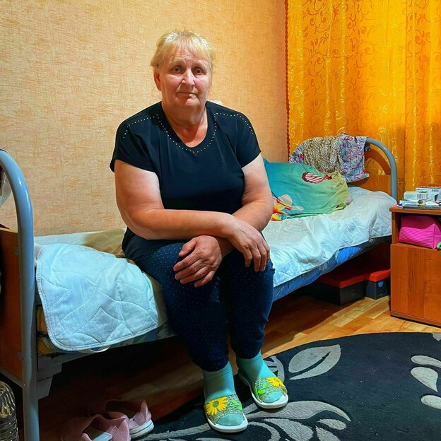 Ljudmilla sitzt auf einem Bett in einem Zimmer mit gelben Vorhängen und gelber Tapete