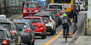 Autos fahren dicht an dicht über eine Brücke, zwei Fahrradfahrer*innen fahren auf einem Radstreifen.