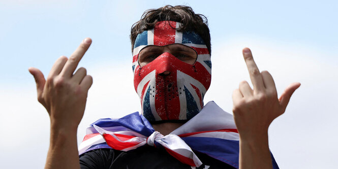 Ein Mann mit Union Jack Maske hält zwei Mittelfinger hoch