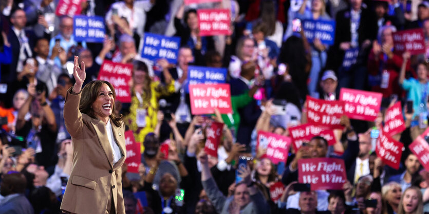 Kamala Harris auf einer Bühne vor jubelnden Menschen mit Schildern, Aufschrift: We Fight, We Win