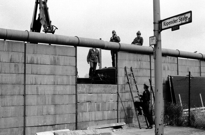 Schwarz-Weiß-Aufnahme, die einen Teil der Berliner Mauer and der Köpenicker Straße zeigt. Drei Soldaten stehen auf der einen, ein Soldat auf der anderen Seite der Mauer. Ein Soldat richtet einen Fotoapparat in Richtung der Fotografin des Bildes.