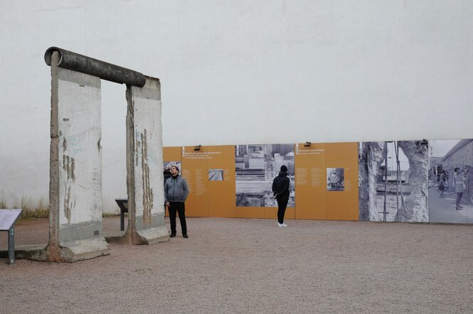 Farbaufnahme mit einem ehemaligen Element der Berliner Mauer. Dahinter sind Tafeln zu sehen, die die Geschichte der Berliner Mauer wiedergeben.