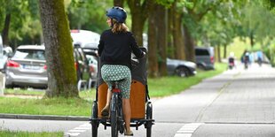 Eine junge Frau mit Helm fährt auf einem Lastenrad auf einem mit Bäumen gesäumten Fahrradweg