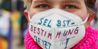 Ein junger Mensch trägt einen pinken Schal und darüber eine FFP-2-Maske mit der handschriftlichen Aufschrift "Selbstbestimmung jetzt" in den Farben der trans Flagge, blau und pink