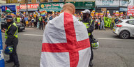 Eine Person steht in eine Englandfahne gewandt auf einer Straße, im Hintergrund demonstrieren andere Personen