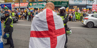 Eine Person steht in eine Englandfahne gewandt auf einer Straße, im Hintergrund demonstrieren andere Personen