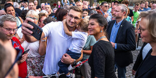 Eine Person in einer Menschenmenge trägt ein Baby auf seinem Arm und macht ein Selfie mit Sarah Wagenknecht