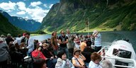 Passagiere sitzen auf Stühlen auf dem offenen Deck eines Schiffes, das durch einen Fjord fährt