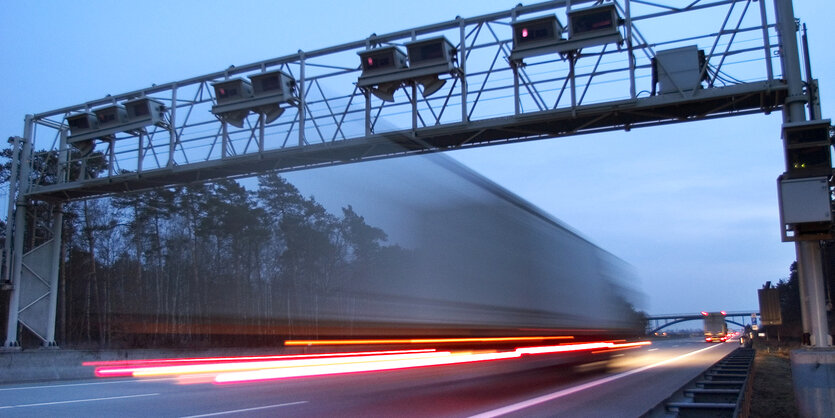 Ein LKW fährt in der Dämmerung unter einer Mautbrückee, die Lichter des LKWS zaubern einen roten Streifen auf die Straße