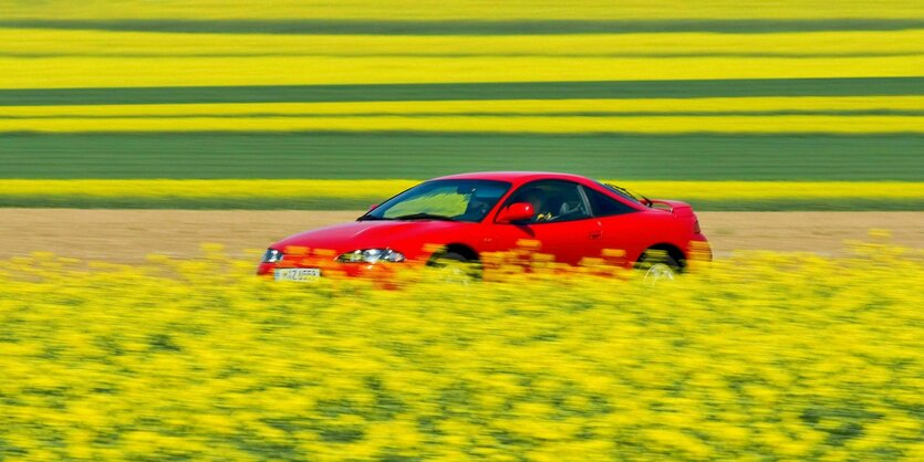 Roter Sportwagen fährt auf einer kleinen Straße durch gelbe Rapsfelder