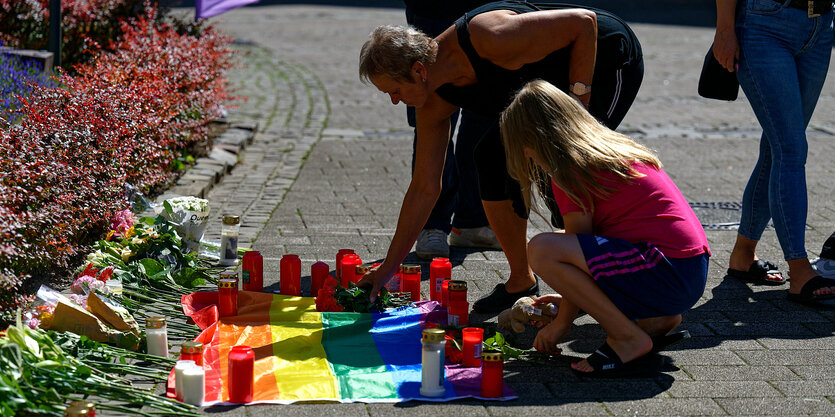 Eine Frau und ein Mädchen legen Blumen auf einer Regenbogenflagge ab. Diese ist mit Kerzen umrandet