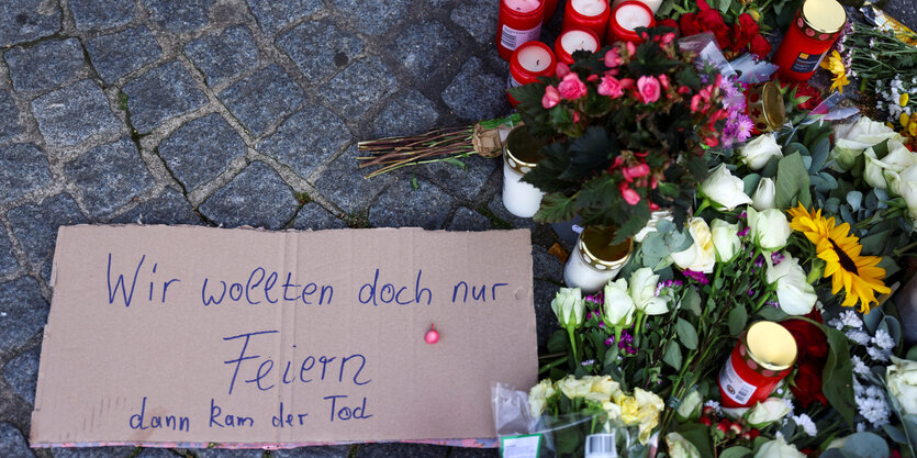 Ein Schild liegt auf dem Boden, auf dem steht "Wir wollten doch nur feiern, dann kam der Tod". Daneben liegen Blumen und Kerzen.