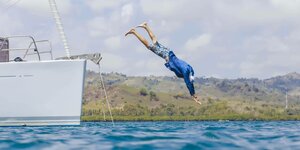 Ein Mann springt in blauer Badehose und Jacke von einer Jacht ins Meer