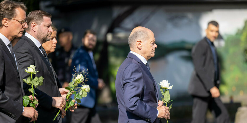 Olaf Scholz mit einer weißen Rose, hinter ihm Hendrik Wüst und andere Regierungsvertreter