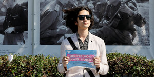 Ein junger Mann mit Sonnenbrille steht mit einem Schild in der Hand vor einer Hecke. Im hintergrund sind historische Fotos zu sehen. auf dem Schild die Forderung: Liberté pour Pavel Dubrow