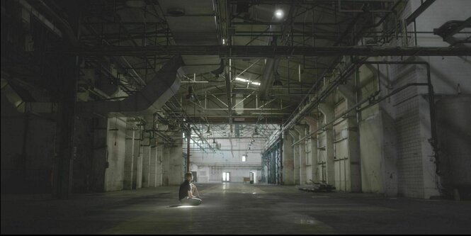 Ein junger Mann hockt in einer großen Industiehalle auf dem Boden.