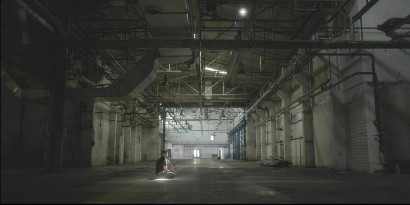 Ein junger Mann hockt in einer großen Industiehalle auf dem Boden.