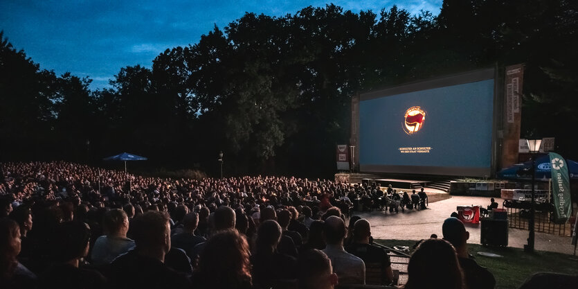 Menschen sitzen in einem Freiluftkino, auf der Leinwand ein Antifa Lobo