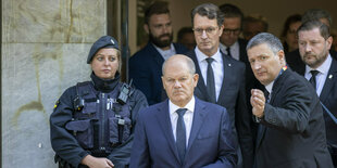 Olaf Scholz mit ernstem Gesicht, hinter ihm steht Hendrik Wüst - an der Seite steht eine schwer bewaffnete Polizistin