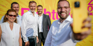 Eine Person macht mit seinem Smartphone ein Selfie mit Christian Lindner und anderen Personen vor einer gelben FDP-Plakatwand