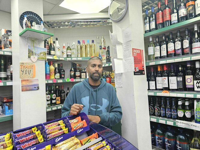 Chanaka Balusurja steht in einem Laden vor enem Flaschenregal, im Vordergrund liegen Schokorigel