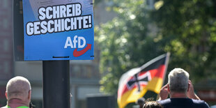 Ein Wahlplakat der AfD mit der Aufschrift «Schreib Geschichte!» hängt an einer Laterne - im Hintergrund ist eine Deutschlandfahne mit Eisernem Kreuz zu sehen