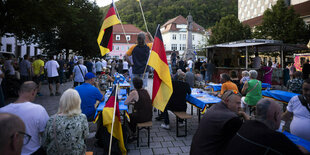 Eine Gruppe von Menschen sietzt auf einem Platz im Freien auf Bierbänken und schwenkt Deutschlandfahnen