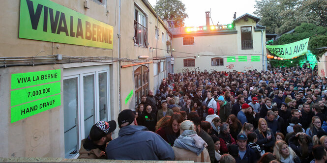 Viele Menschen versammeln sich dicht gedrängt in einem Innenhof, an dessen Mauer "Vive la Bernie" steht.