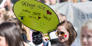 Eine Frau auf einer Demo hält ein Schild hoch, auf dem steht: Pflege mit Herz sollte mehr wert sein