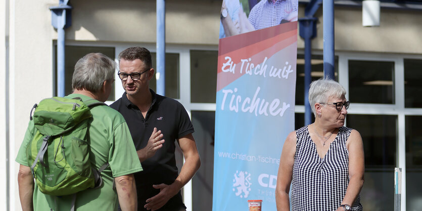 Vor einem Plakat mit der Aufschrift "Zu Tisch mit Tischner" stehen drei Personen. Ein Mann im schwarzen Ti-Shirt diskutiert mit einem mann in grüner Kleidung und mit grünem Rucksack. Eine Frau mit kurzen grauen Haaren steht daneben und schaut von den beiden Männern weg