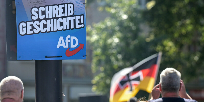 Ein Wahlplakat der AfD und im Hintergrund eine Deutschlandfahne mit eisernem Kreuz.