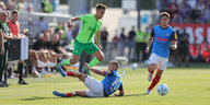 ein Fußballer in blauen Trikot mit der Nummer 10 rutscht auf dem Rasen, einer im grünen Trikot springt über ihn; der Ball ist schon weg