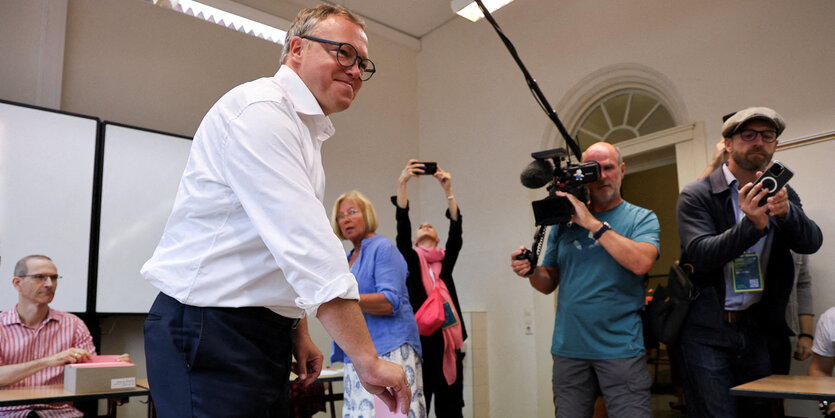 Mann mit Brille wirft grinsend einen Wahlzettel in die Urne