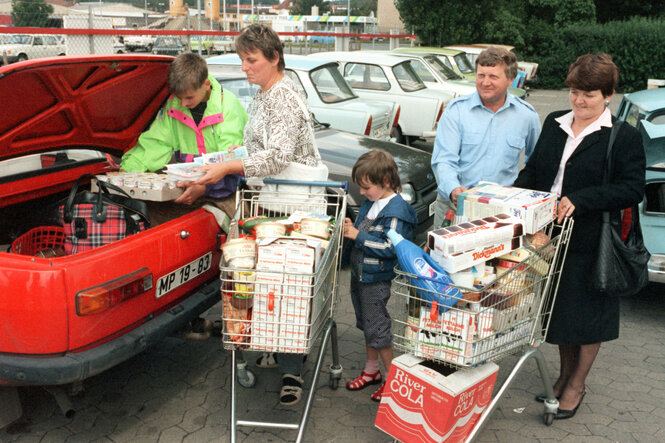 Ein historisches farbfoto aus dem Juli 1990 zeigt DDR-Bürger, die Westprodukte aus vollgepackten Einkaufswagen in den Kofferraum eines roten Wartburgs laden