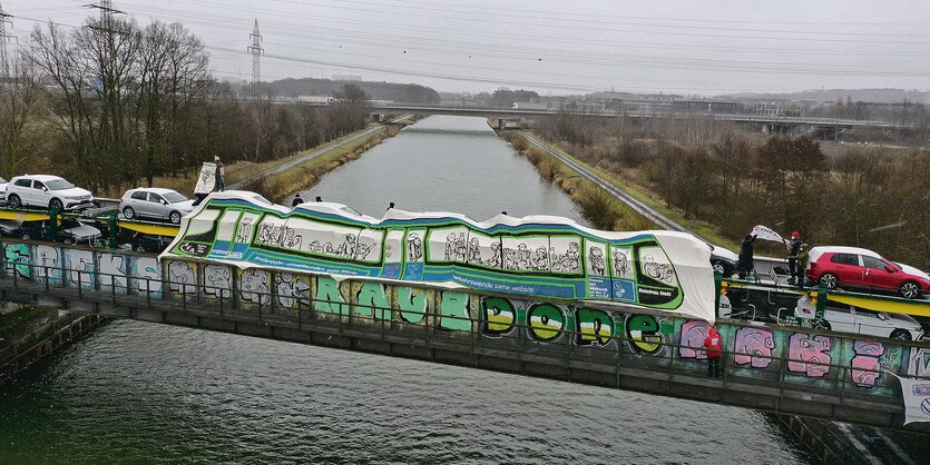 Aktivist*innen verkleiden einen VW-Autozug über dem Mittellandkanal mit einem großen Transparent. Auf dem ist eine Straßenbahn aufgemalt.