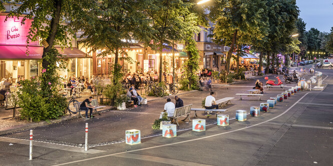 Bemalte Poller grenzen eine Straße ab, dahinter sitzen Mensch auf Bänken