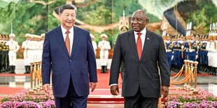 Xi Jinping und Cyril Ramaphosa stehen gemeinsam lächelnd vor einer Kulisse aus kitschigem Bild der chinesischen Mauer und aufgereihtem Militär