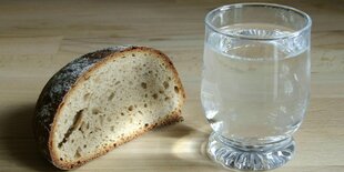 Ein halbes kleines Brot und ein Glas Wasser stehen auf einem Tisch