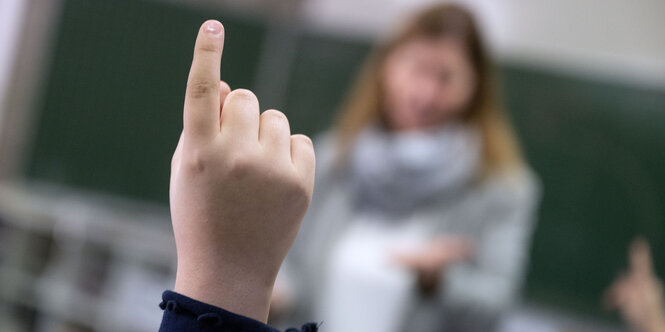 Eine Kinderhand ragt ins Bild, ein Schüler meldet sich, im Hintergrund ist verschwommen eine Lehrerin vor einer Tafel zu erkennen