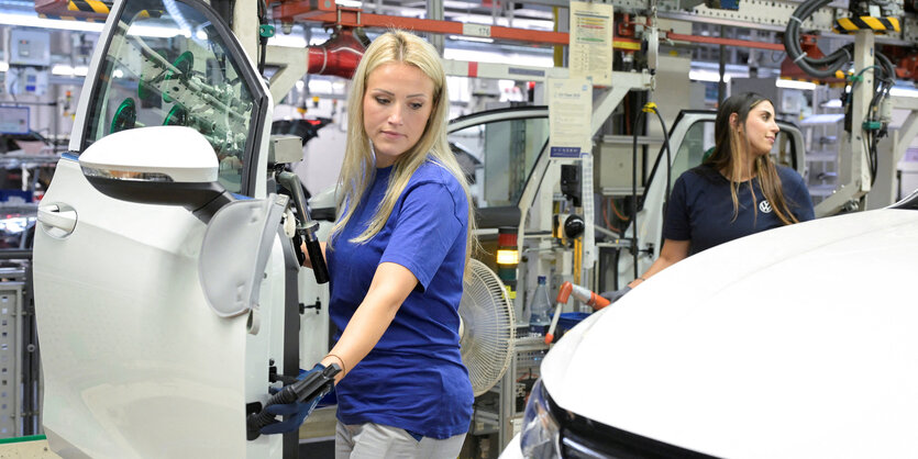 Zwei junge Frauen in der Montagehalle von VW, eine junge Frau beweght eine weisse Autotür Richtung Golf III