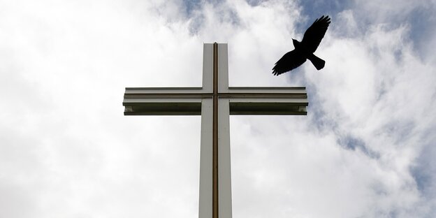 Vor einem bewölkten Himmel steht ein Kreuz um den ein schwarzer Vogel fliegt