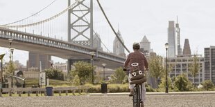 Ein Mann fährt mit dem Fahrrad auf das Flussufer einer Stadt zu. Dahhinter ist eine große geschwungene Brücke zu sehn. Auf dem Gepäckträger seines Fahrrads befindet sich ein Kackhaufen mit aufgesetzten Scherzartikelaugen.