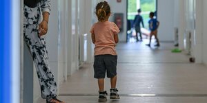 Ein Kind sgteht im Flur einer Asylbewerberunterkunft und schaut anderen Kindern beim spielen zu. Eine weibliche Person tritt aus der Tür, ist aber nur halb zu sehen