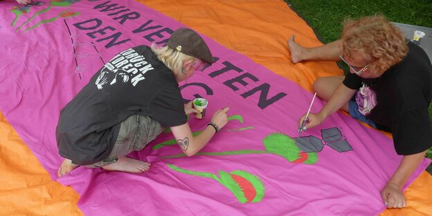 Zwei Frauen bemalen auf dem Boden sitzend ein Transparent, auf dem Pflanzen Bomben fressen