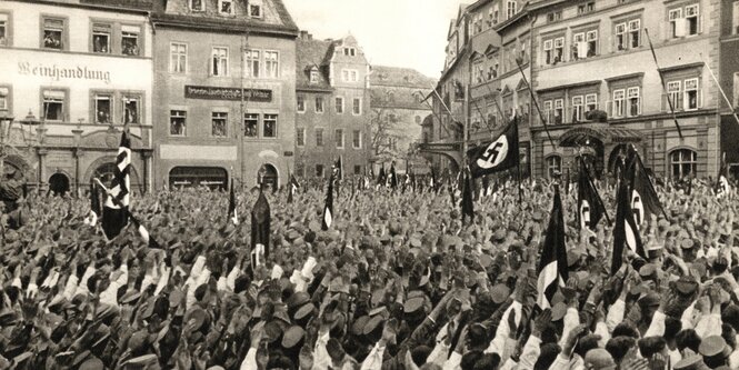 Ein historisches Foto zeigt eine große Menschenansammlung auf einem Marktplatz. Einige Personen schwenken Fahnen mit Hakenkeuzmotiv.