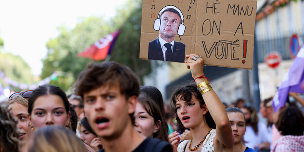 Mehrere junge Menschen auf der Straße, die protestieren. Eine Frau hält ein Schild hoch, auf dem Macron abgebildet ist - mit Kopfhörern. Auf dem Schild steht: He, Manu, wir haben gewählt.