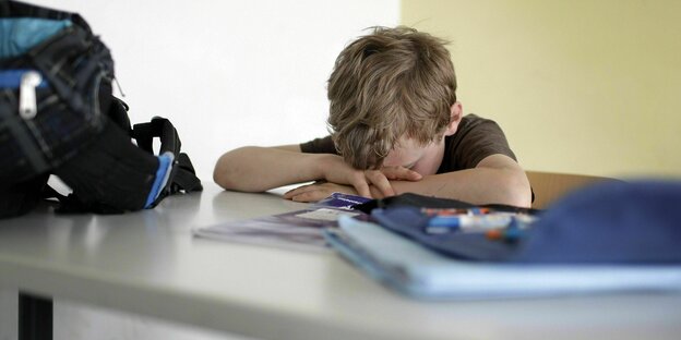 A sad child with his head resting on his arms sits at a classroom table, his bag and notebooks next to him